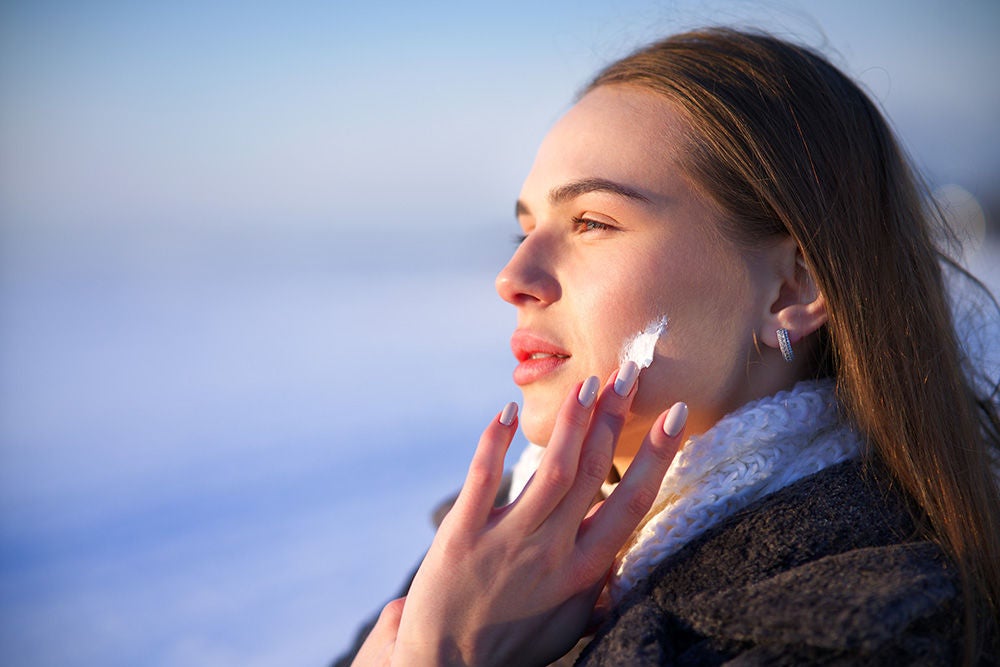 Portrait of beautiful woman applying cosmetic skin cream on face and hands, protecting from cold weather at winter season. Pretty girl take care of her skin outdoors on snowy nature background. 